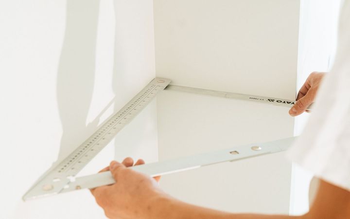 Hands holding a metal square against a white wall corner