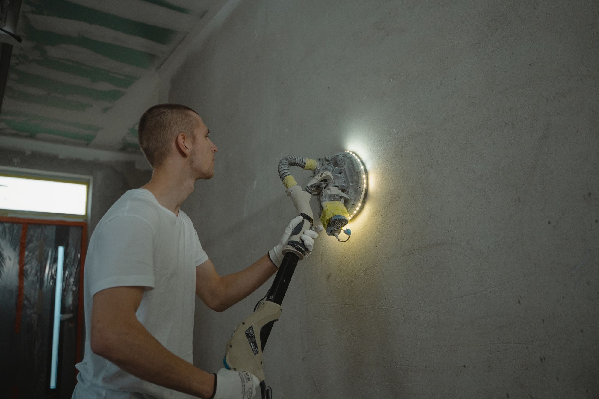 Worker sanding a bare interior wall with a pole sander and a bright light