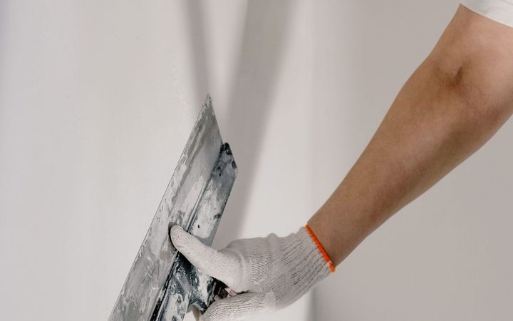 Gloved hand drawing a putty knife across a white wall