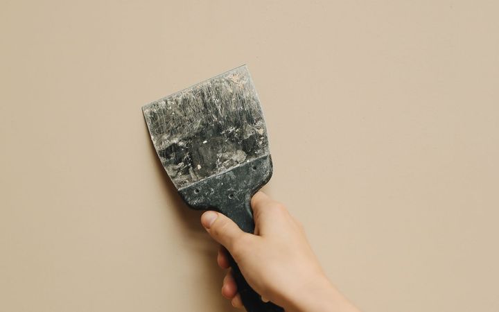 Hand pressing a putty knife against a beige painted wall